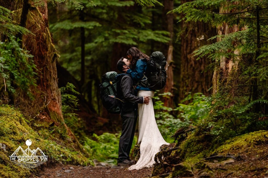 A bride and groom kiss in the forest with backpacks during their Washington elopement.