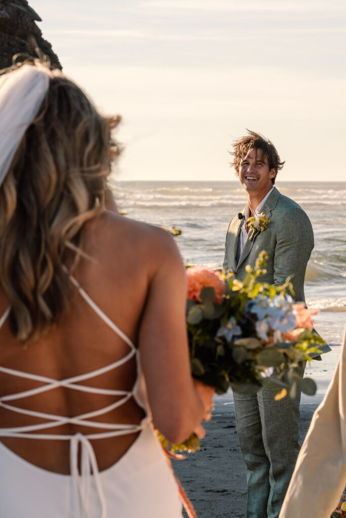 A bride walks down the aislee during her coastal elopement as their oregon coast elopement photographers shoot over her shoulder to capture her grooms reaction.