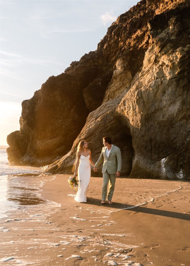 A bride and groom explore a rocky beach during the sunset of their coastal elopement. 