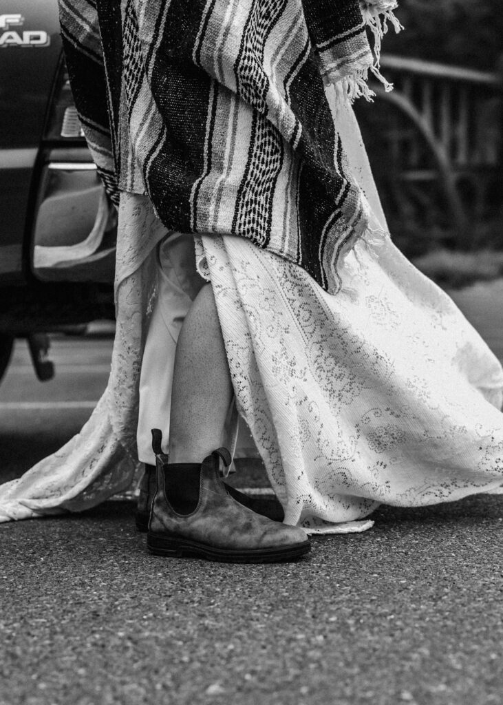 A detail shot focusing on the footwear of the bride from this surfer wedding. her dress billows over her Blundstone boots, and the ends of her camp blanket hang near her ankles