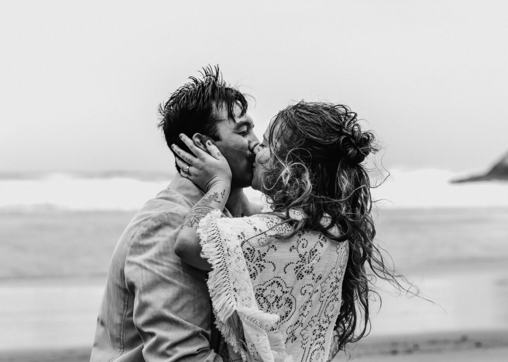 A black and white image of a bride and groom sharing their first kiss on a moody beach during their surfer elopement.