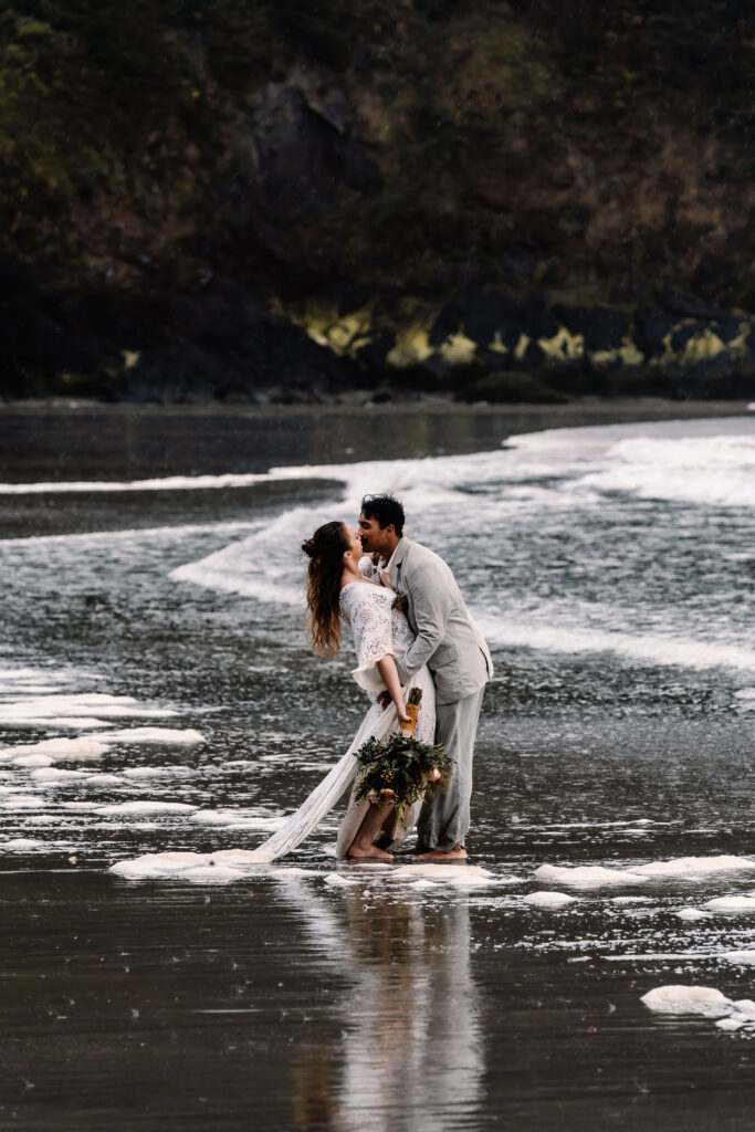 a bride and groom kiss on a moody beach following their surfer wedding. The waves roll over their bare feet.