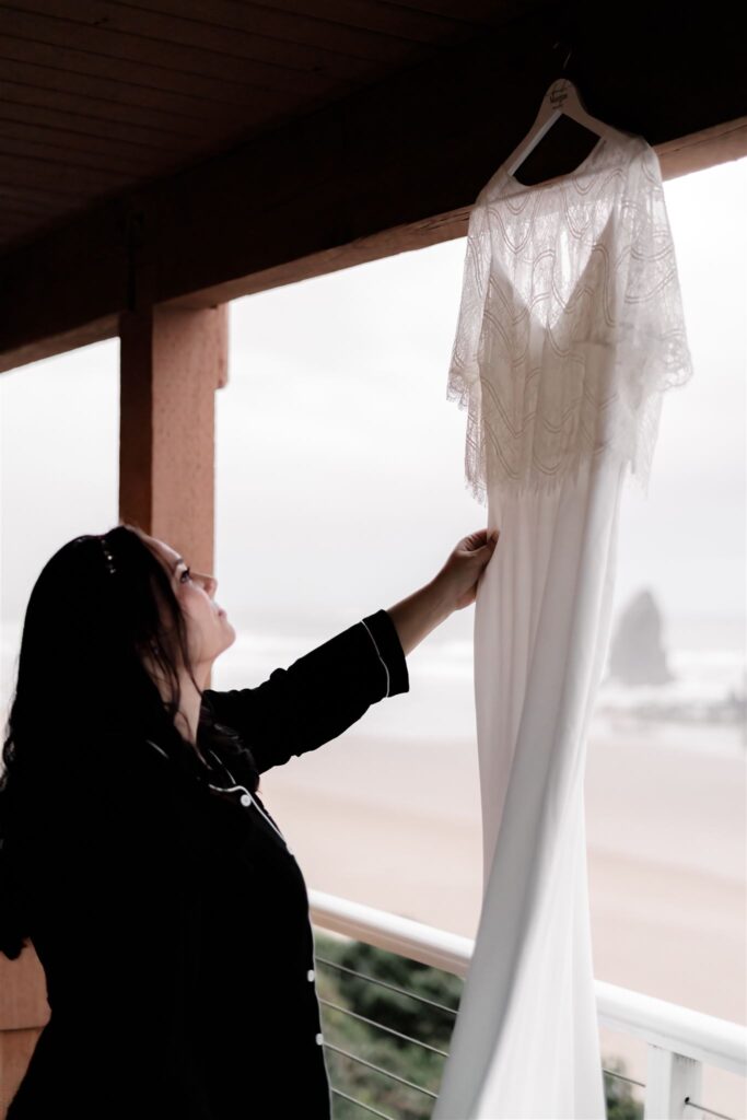 as she prepares for her Pacific Coast wedding, a bride gazes at her wedding dress that hangs outside. The silhouette of a foggy, rocky beach can be seen in the background. 