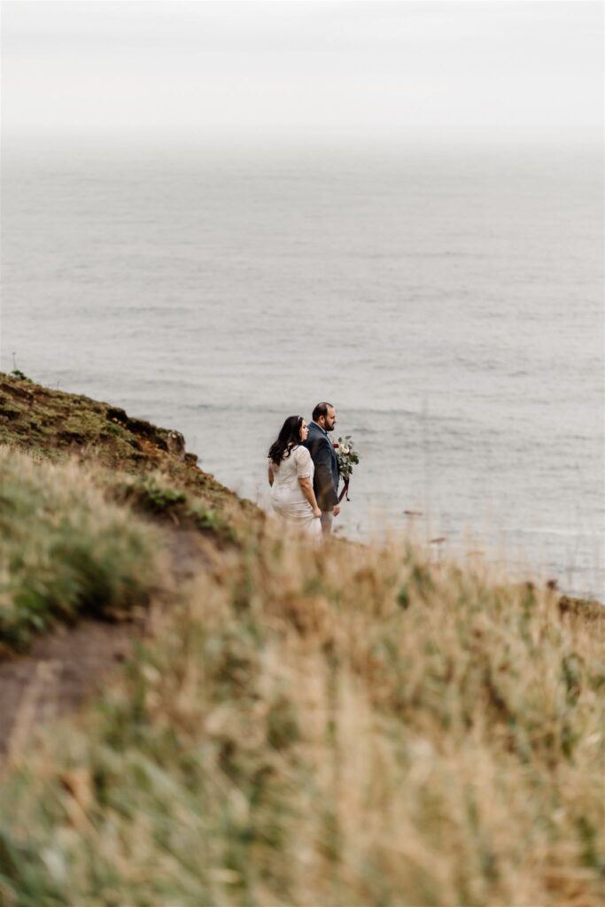 framed by green grass, a couple in wedding attire explore the ocean cliffside during their Pacific Coast wedding.
