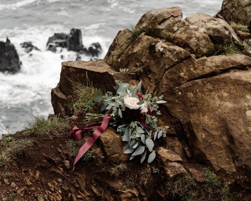 A romantic bouquet of roses and eucalyptus lay delicately on a rocky seaside cliff. The bride has laid them down as she explores during her Pacific Coast Wedding. The wine colored, silk ribbon billows in the wind. 