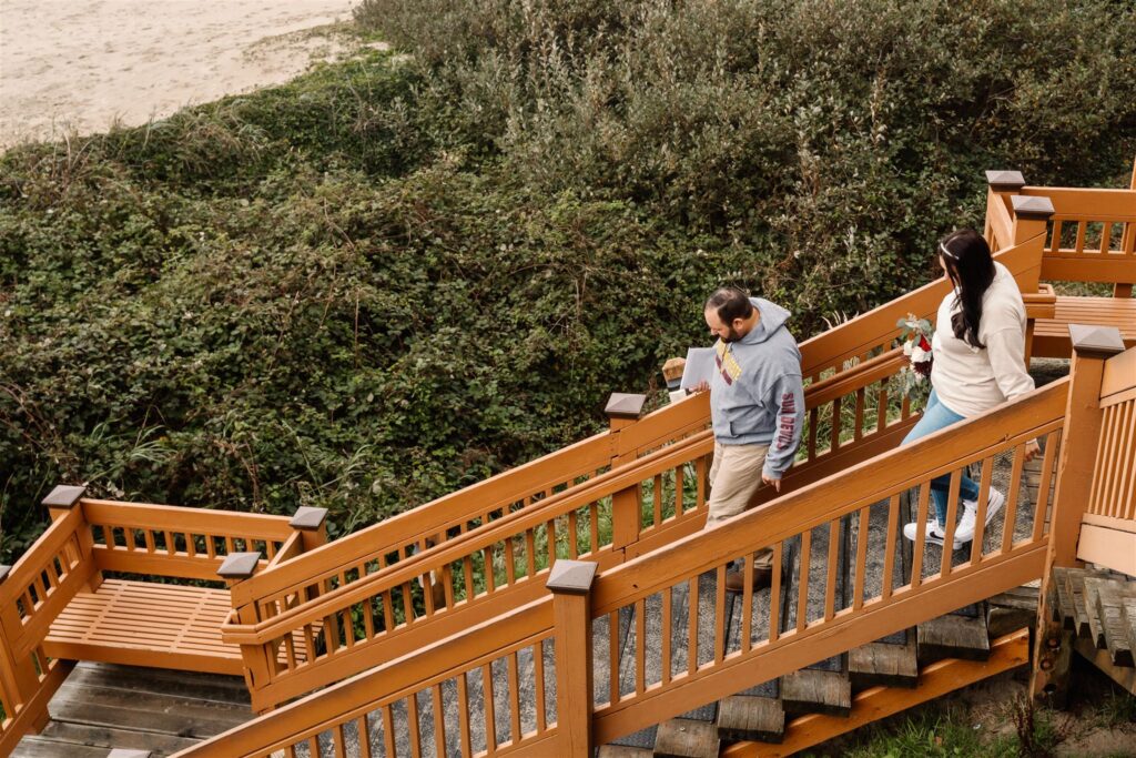 after exploring, a bride and groom in comfortable clothing head down a wooden staircase to their beach reception to celebrate their pacific coast wedding
