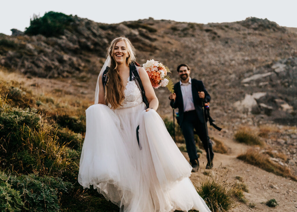 a bride smiles widely as she hikes in her wedding attire, her groom following behind her