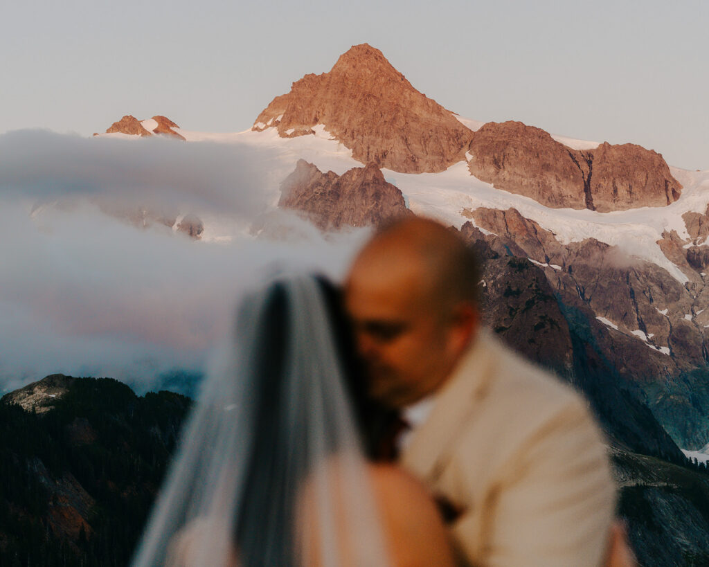 a sharp image of the mountains drenched in pink alpine glow. A couple embraces in the foreground during this last minute elopement