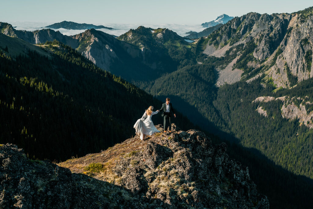 a massive mountain landscape illuminated by the sun. On a cliff highlighted by golden sunshine we see a couple moving to the center. They are exploring the landscape around them during their Olympic Peninsula elopement