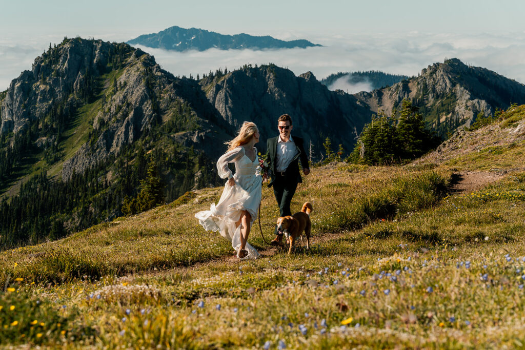 a couple in wedding attire runs through a field in their wedding attire with mountains in the background during their Olympic Peninsula elopement