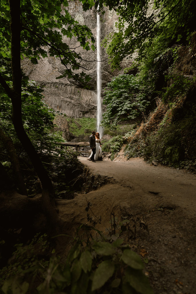 Couple in wedding attire walks through forest with plunging waterfall in the background during their waterfall elopement
