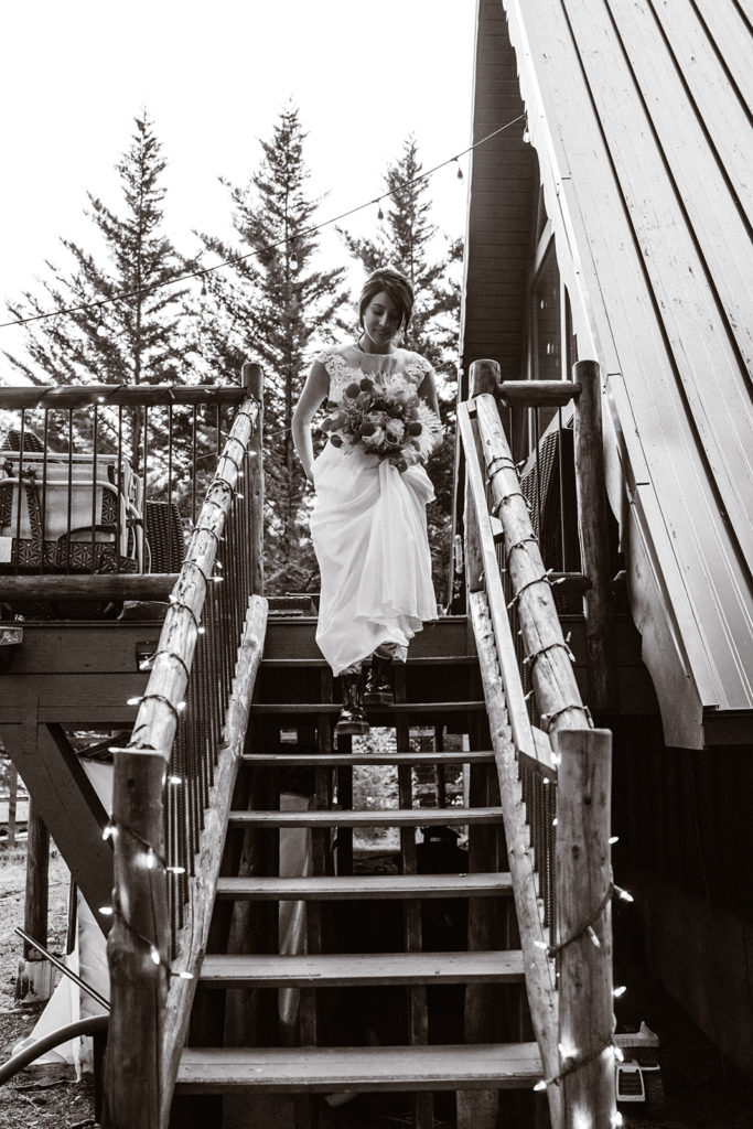 black and white photo of a bride walking down the steps of their aframe cabin holding her boquet during her mt. rainier wedding