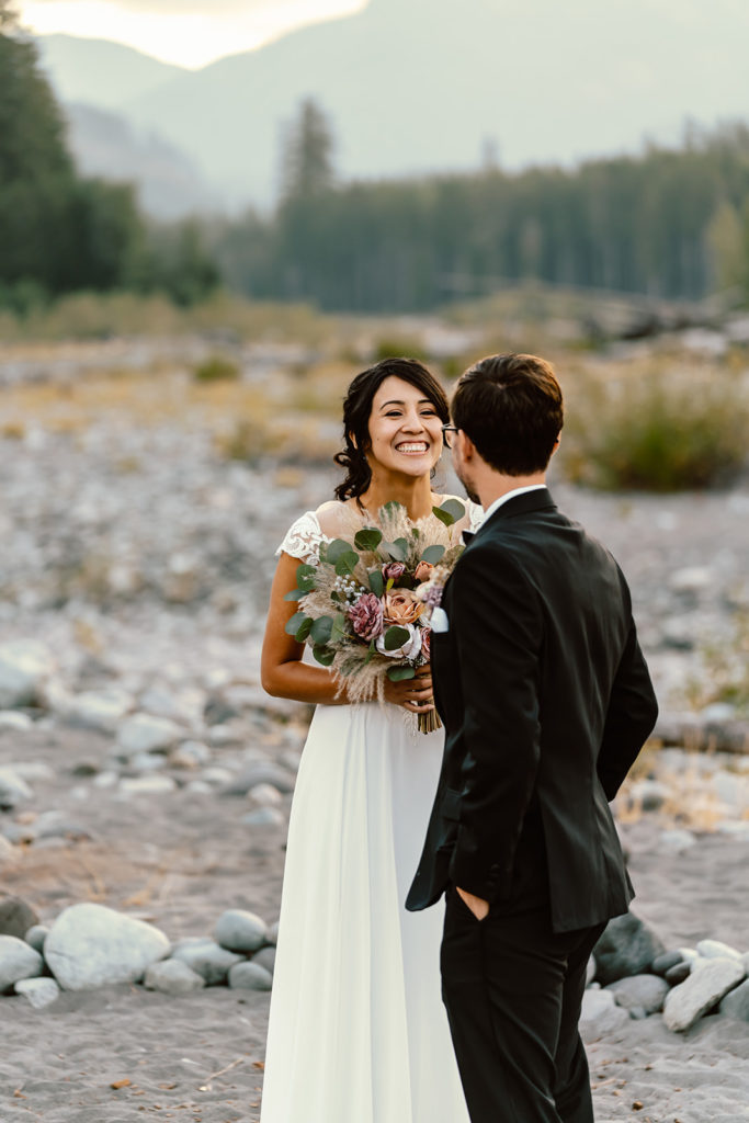 Bride and groom in their wedding attire smiling as they share a first look near the river during their mt rainier wedding