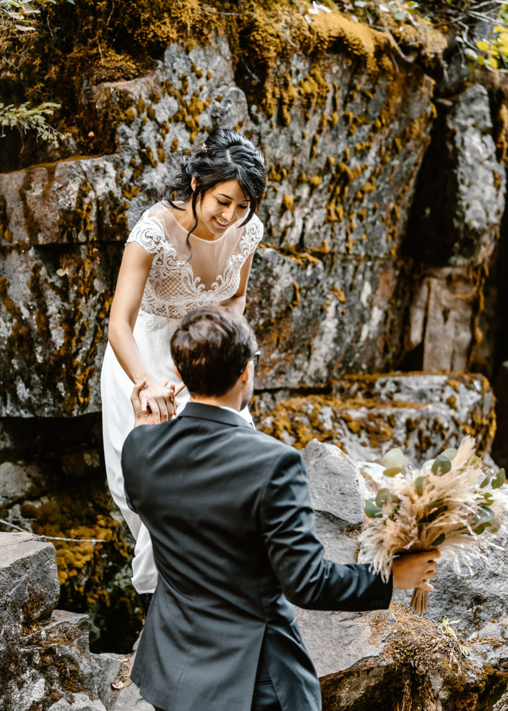 Groom holds bouquet as he helps his bride climb down to their mt. rainier wedding location