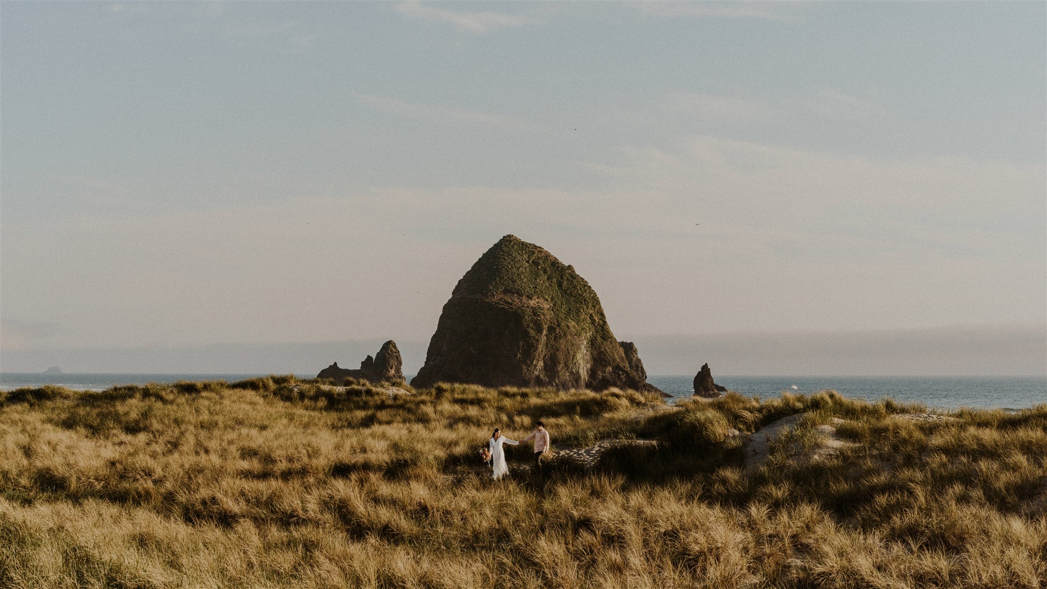 After considering elopement vs wedding, a couple in wedding attire walks through seagrass and sand dunes in front of Haystack rock during their elopement.