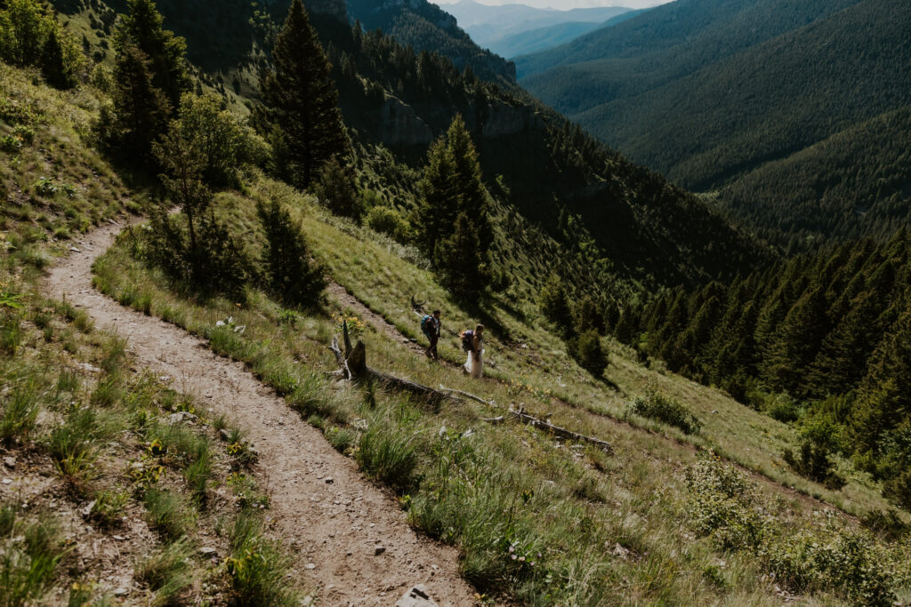 After considering elopement vs wedding, a couple in wedding attire follows a curving switchback with their camping packs. A stunning forested canyon and blue-green mountain range serves as the backdrop to their elopement day.