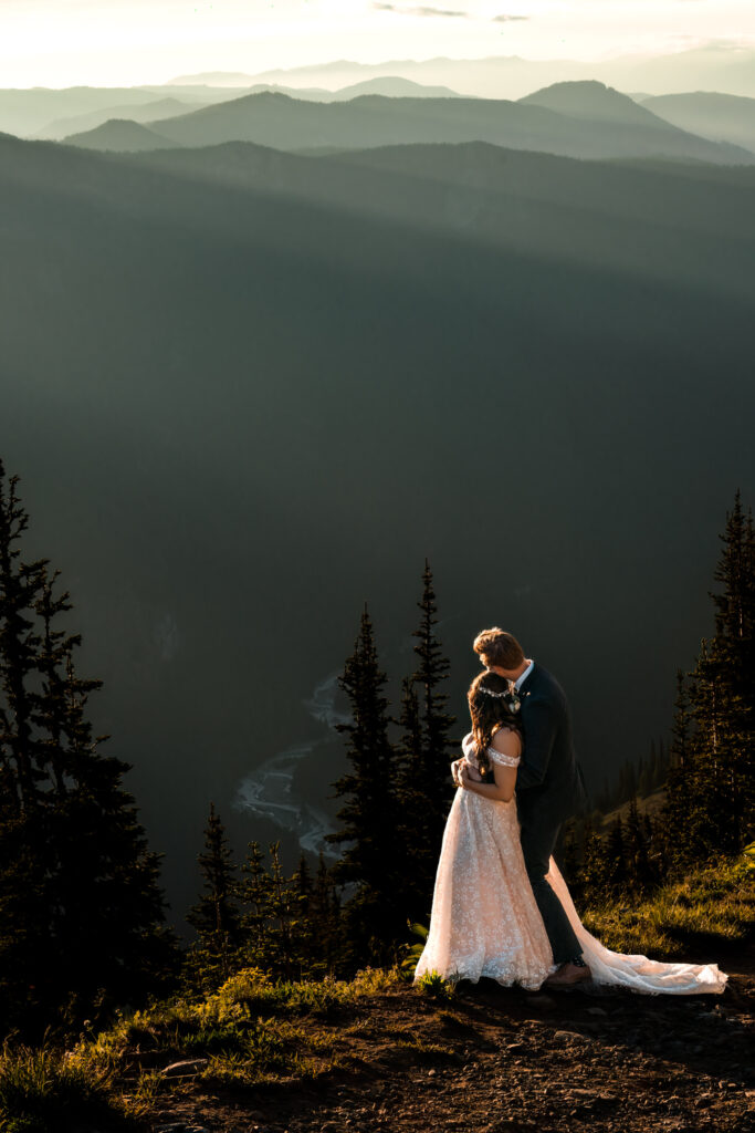 After considering elopement vs wedding, a couple in wedding attire gazes out at a the sweeping mountain views as the sun sets, painting the mountain range silhouettes in layers and ranges of blue, teal, navy and gold. A river lays below them and they are framed by growing pines. 