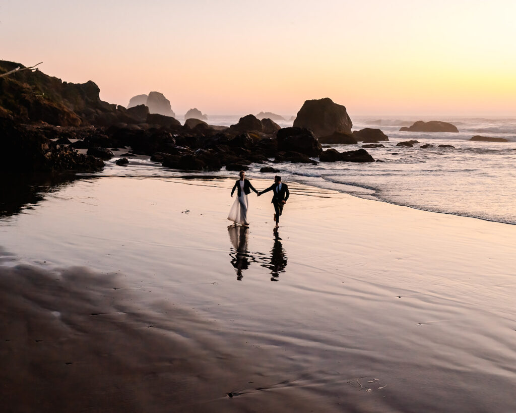 After considering elopement vs wedding, a couple in wedding attire run down a rocky coastline with jagged sea stacks and rocks jut out from the sand