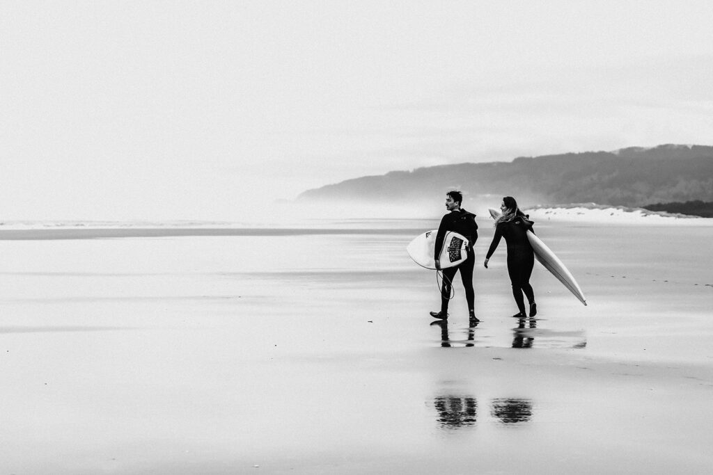 After considering elopement vs wedding, a couple in wetsuits walk along an empty beach towards the ocean carrying their surfboards towards the water as part of their elopement day