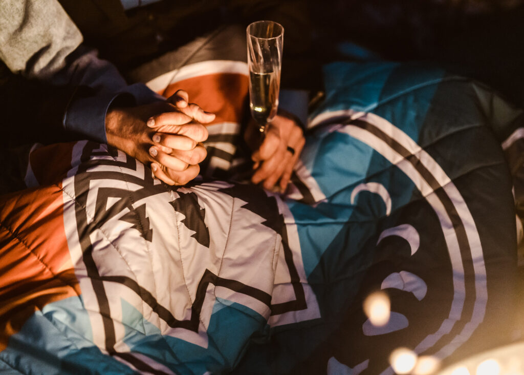 At the end of their elopement timeline a couple holds hands by a campfire, one hand holding champagne, the other embraces the hand of their partner 