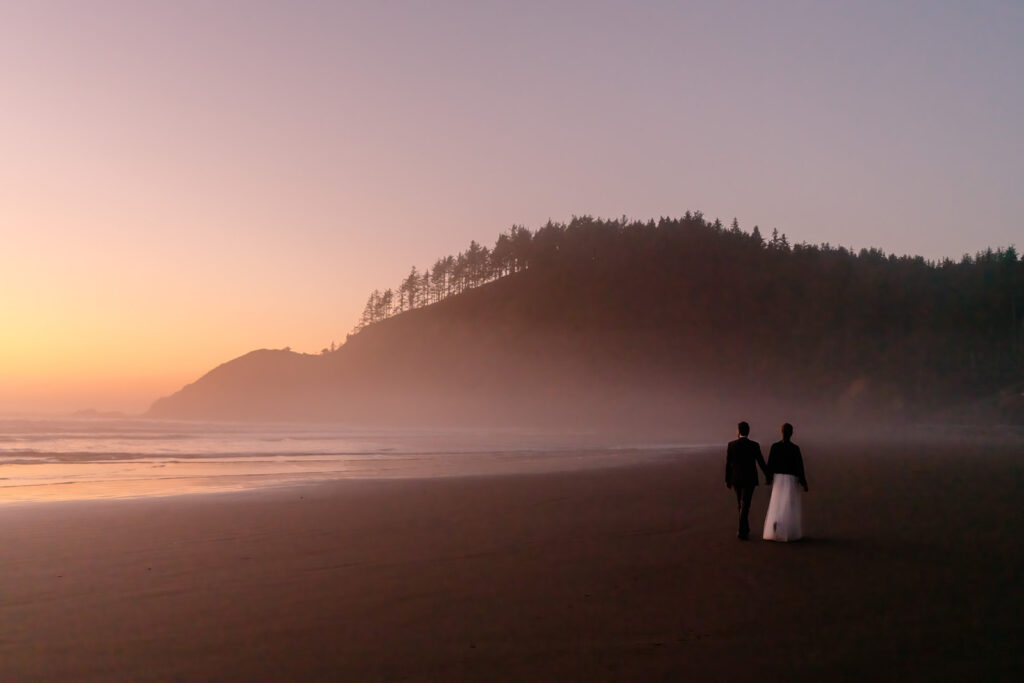 at the end of their elopement timeline, a couple in wedding attire walks along a foggy, rocky beach. The sun sets behind the Pacific casting a fleeting, pink and gold light on the gentle waves.