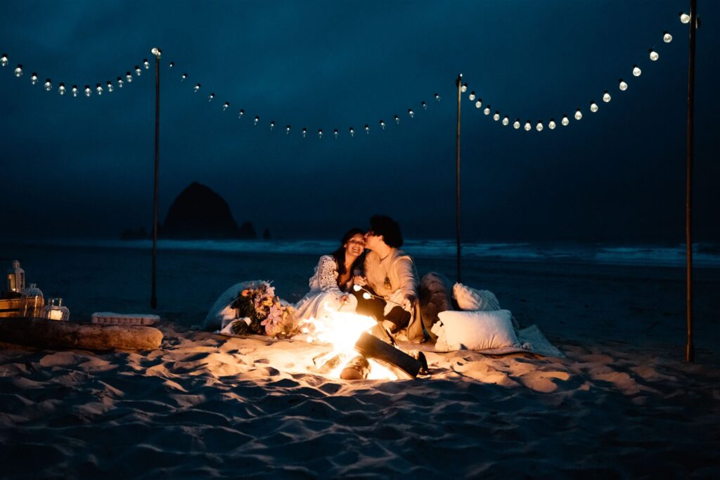 A couple ends their elopement timeline snuggling in front of a large haystack rock by a small beach fire. 