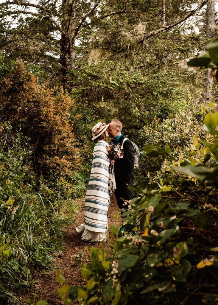 A couple stops to kiss in a lush green forest in wedding attire holding a bright bouquet of dried flowers. The bride wears a hat with a flower crown and wraps herself in a blanket 