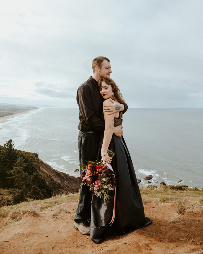 A couple in all black wedding attire embrace at the top of a green cliff. The blue waves of the pacific can be seen rolling beneath them. 