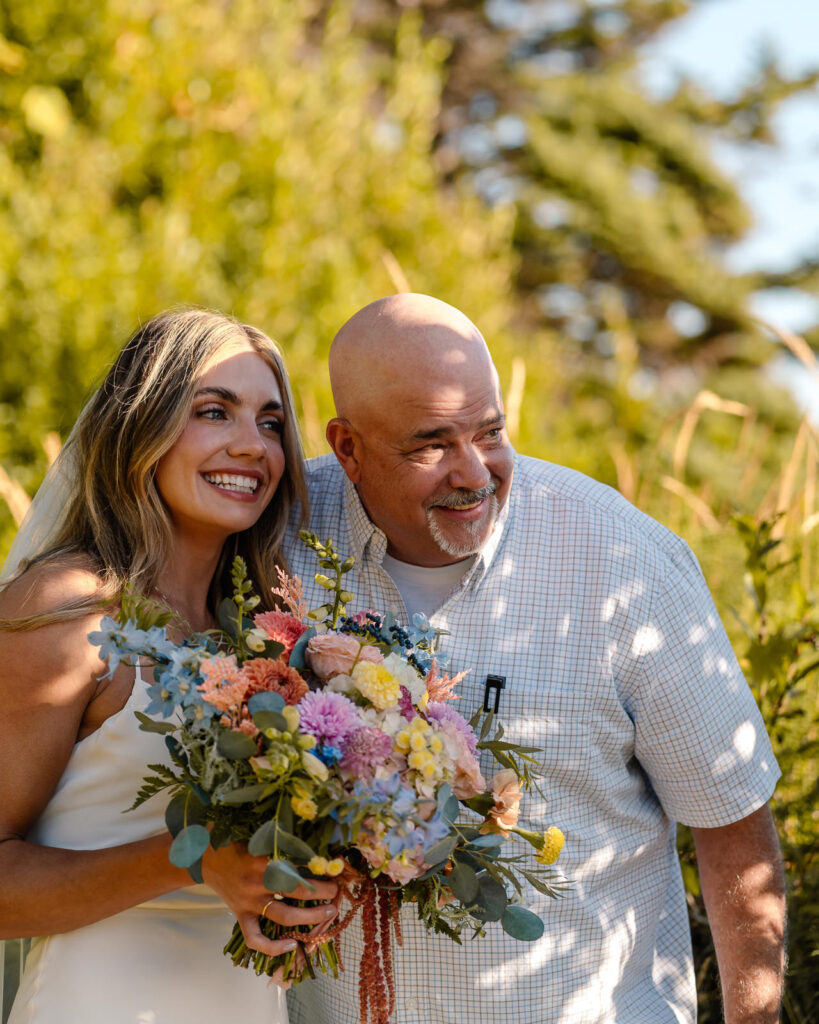 A bride waits with her father for the ceremony of her Oregon coast elopement. They grin at the crowd while peeking behind bushes 