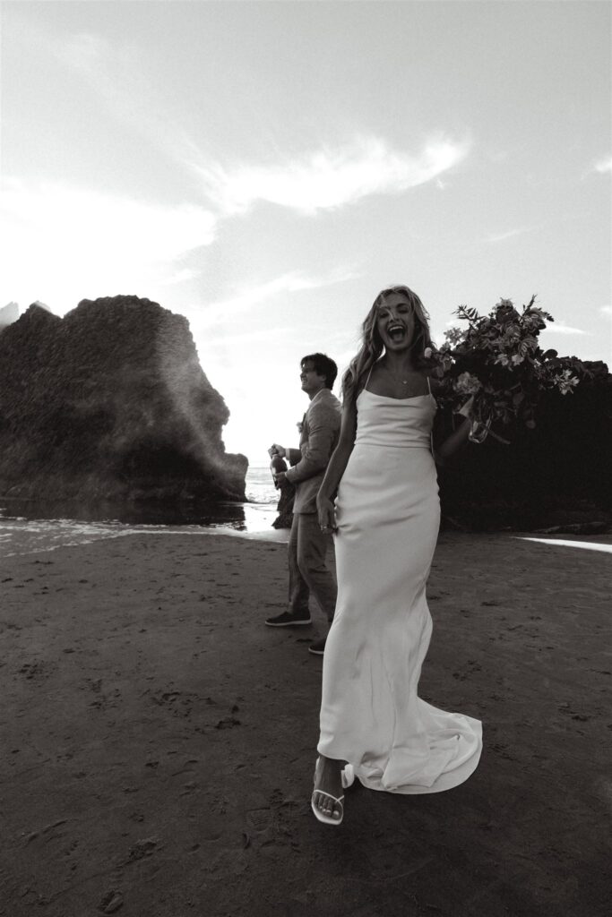 a bride smiles widely at the camera as her groom pops a bottle of champagne to celebrate their coastal elopement