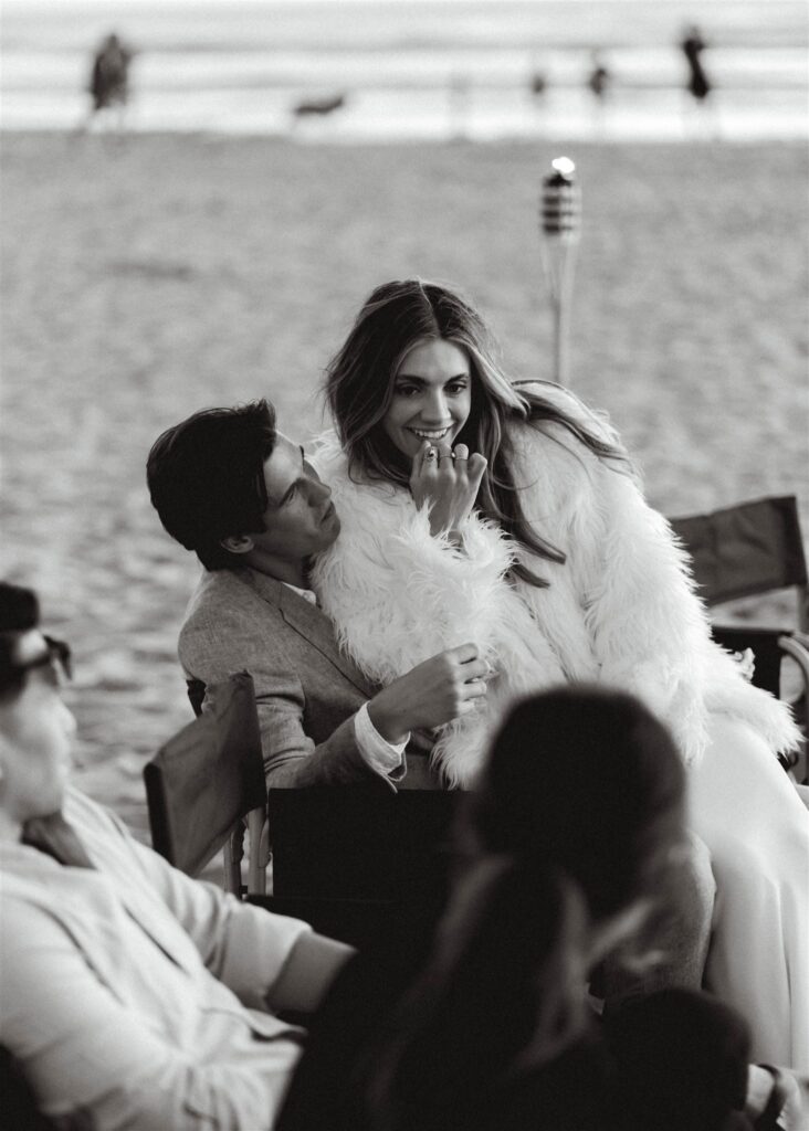 During the bonfire beach reception of their coastal elopement, a bride and groom share a chair and beer to celebrate their day. 