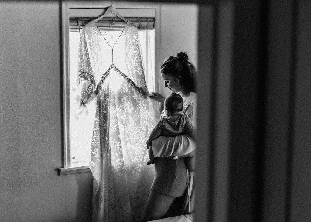 A bride admires her wedding dress with her baby in her hand, as she prepares for her surfer wedding.