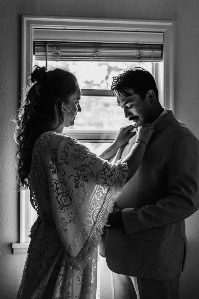 A black and white image of a bride helping a groom get dressed for their surfer wedding.