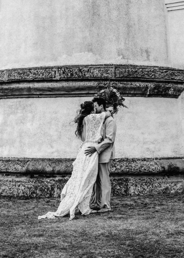 A black and white photo of a couple kissing at the base of a light house in wedding attire. There is wind whipping through their clothes.
