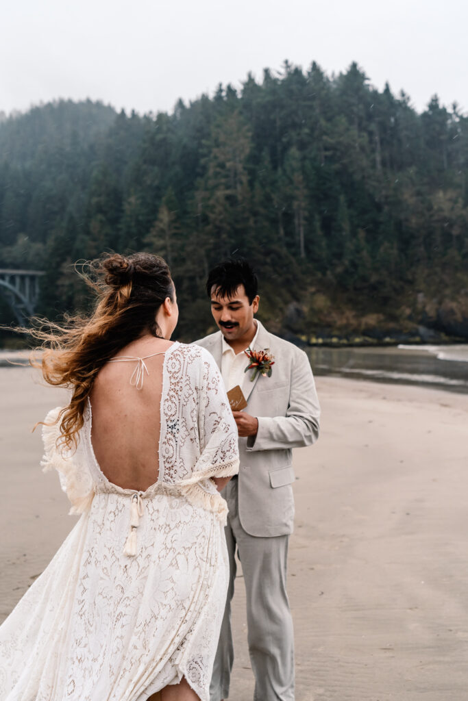 An image of a groom reading his vows to his bride on a moody beach during their surfer wedding