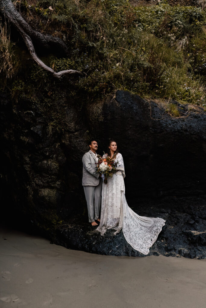 As they explore the beach after their surfer wedding, a couple seeks shelter from the downpour inside a shallow sea cave. roots of a tree above them cascade down the side of the cave as they gaze towards the far out ocean.