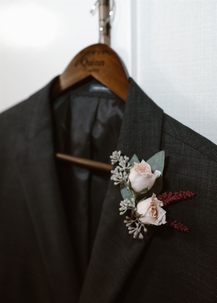 a boutonnière is fastened to the lapel of a grooms suit. Two pale, pink roses nestled with greenery.