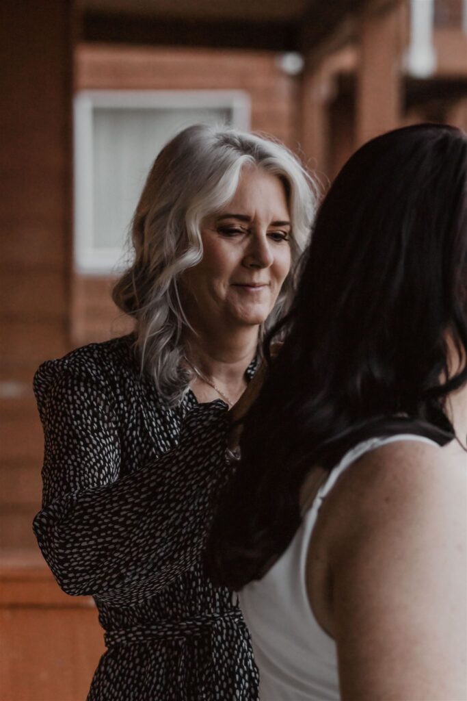 a mother helps her daughter prepare for her pacific coast wedding. She stands to the side of her daughter and helps to secure the straps of her wedding dress.