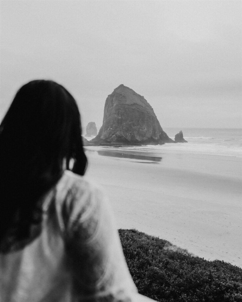 A bride gazes out at a large haystack rock on an empty beach as she gets ready for her Pacific Coast wedding.