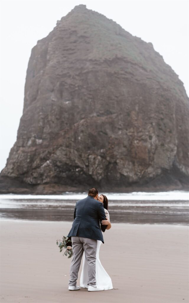 a couple in their wedding attire kiss in front of the iconic haystack rock on cannon, beach. The fog rolls in and creates a moody vibe for their Pacific Coast wedding.
