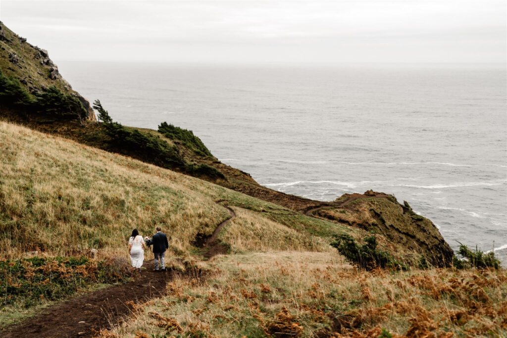 a wide landscape shot of a bride and groom explore a green trail that leads to an open cliffside during their pacific coast wedding