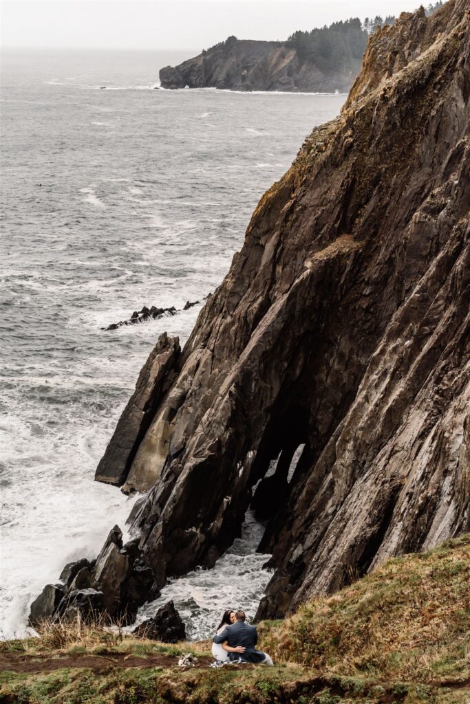 during their Pacific Coast Wedding, a couple in their wedding attire sits on a coastal cliff. They gaze out at the jagged cliffs and ocean below them.