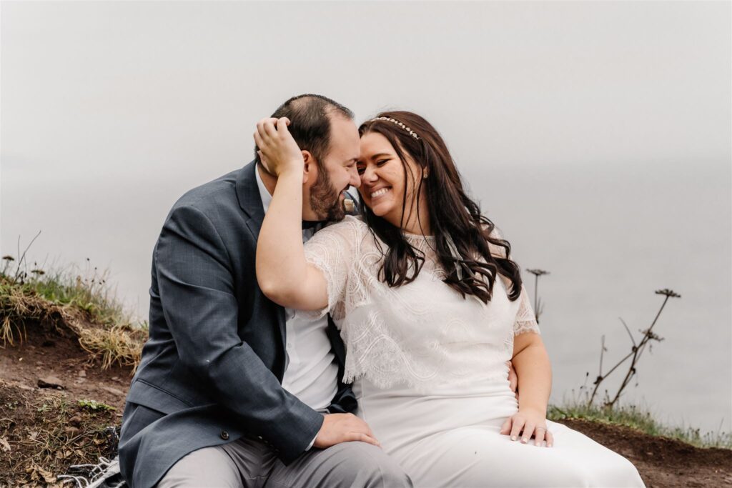 during their pacific coast wedding, a bride and groom snuggle and smile as they sit on a blanket in their wedding attire