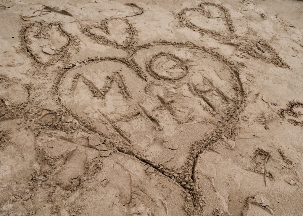 a detail shot of the initials of the bride and groom inside of a heart that is drawn in the sand to commemorate their pacific coast wedding