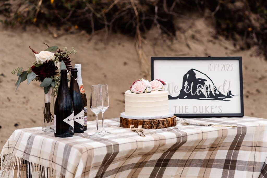 a detail shot of the reception table for this pacific coast wedding. it is decorated with roses, a wedding cake, an elopement sign, and local wine