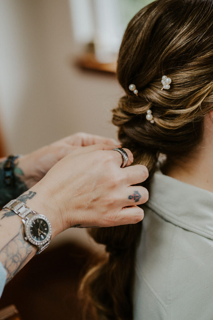 A detail shot of the brides hair and pearls woven into her braid by her skillful makeup artist, whose hands can be seen working.