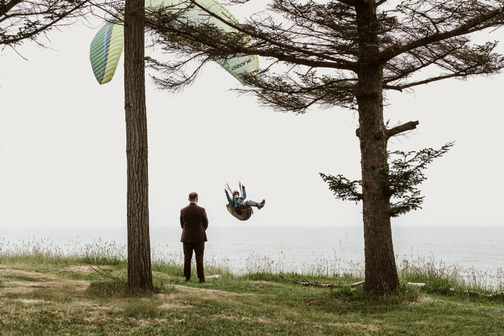 The groom stands between two trees facing away from us. It is clear that he is waiting to see his bride. A man paragliding is in front of him, flying over the sea.