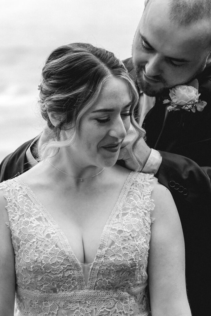 A black and white photo as our bride smiles while her groom stands behind her fastening her necklace. A gift from him to commemorate their forest wedding.