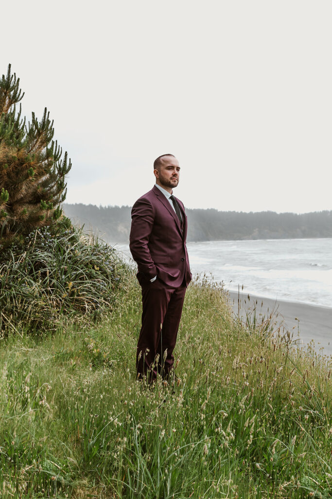 A groom stands cliffside in his maroon suit gazing out at the sea