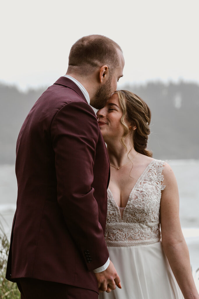 A groom kisses his bride on the forehead as she smiles, their hands are clasped together.